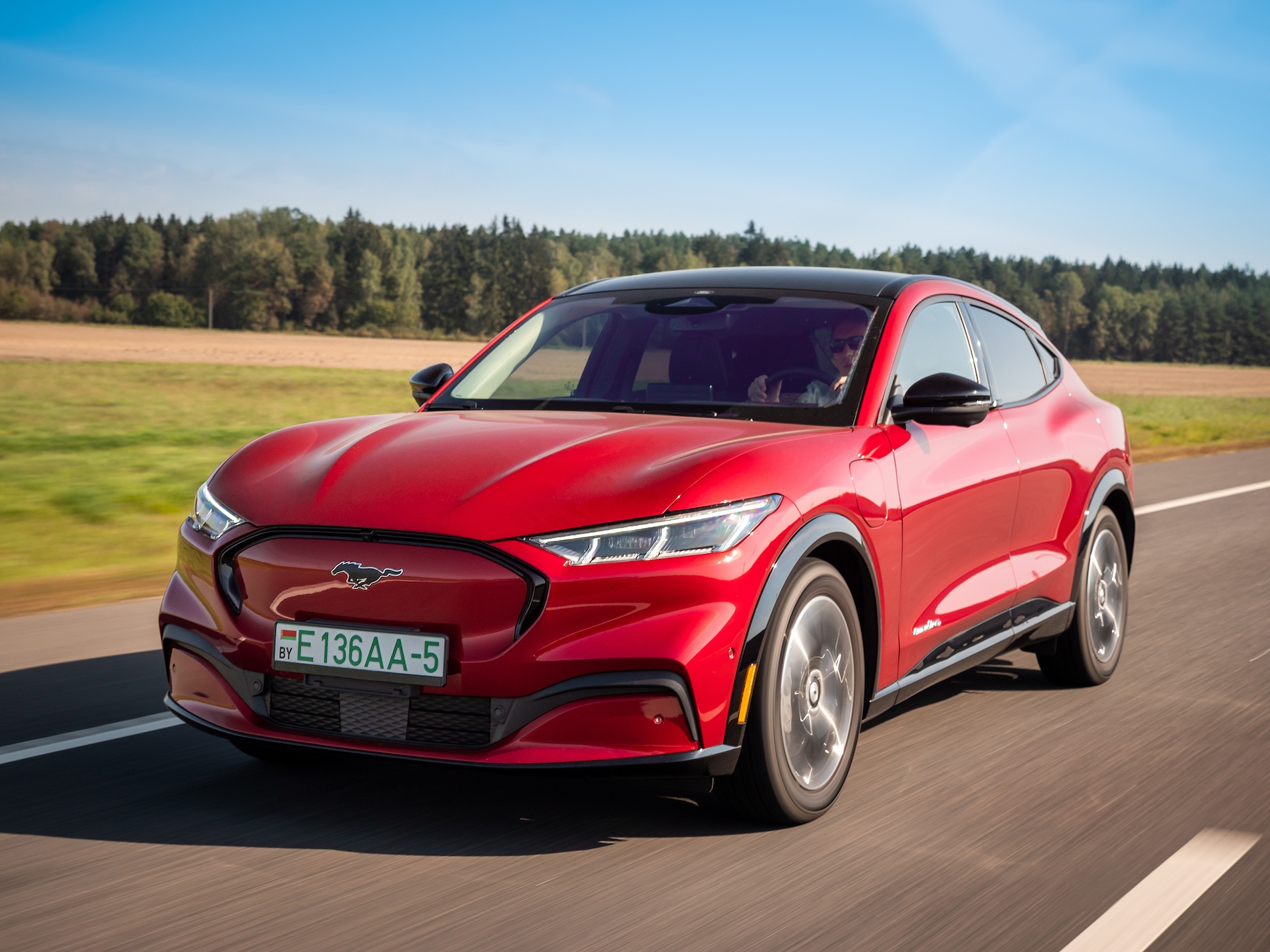 All-electric bright red Ford Mustang Mach-E drives on a highway during a sunny day.